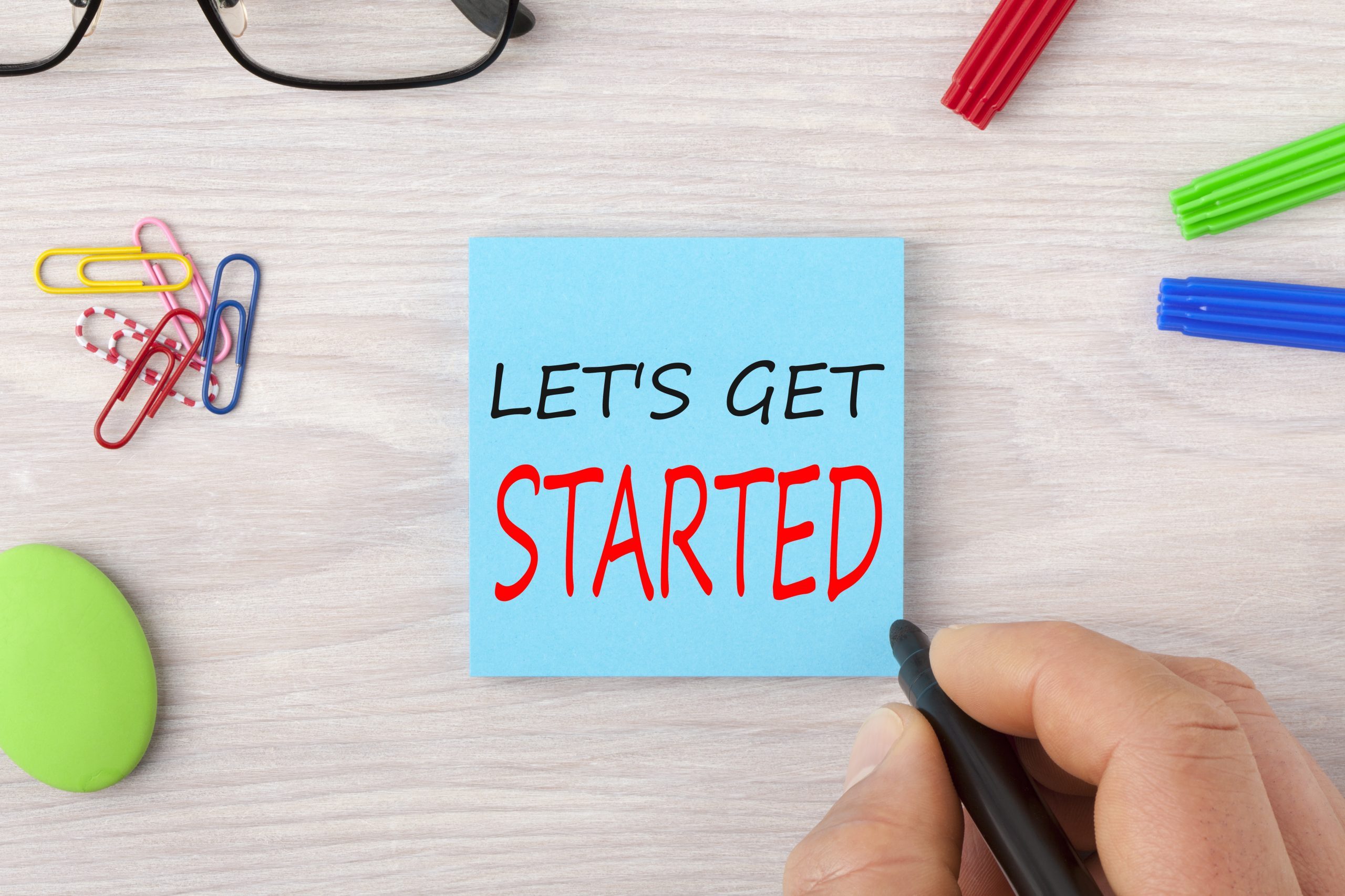 Hand writing LET'S GET STARTED in note with marker pen and glasses on wooden desk. Business Concept.